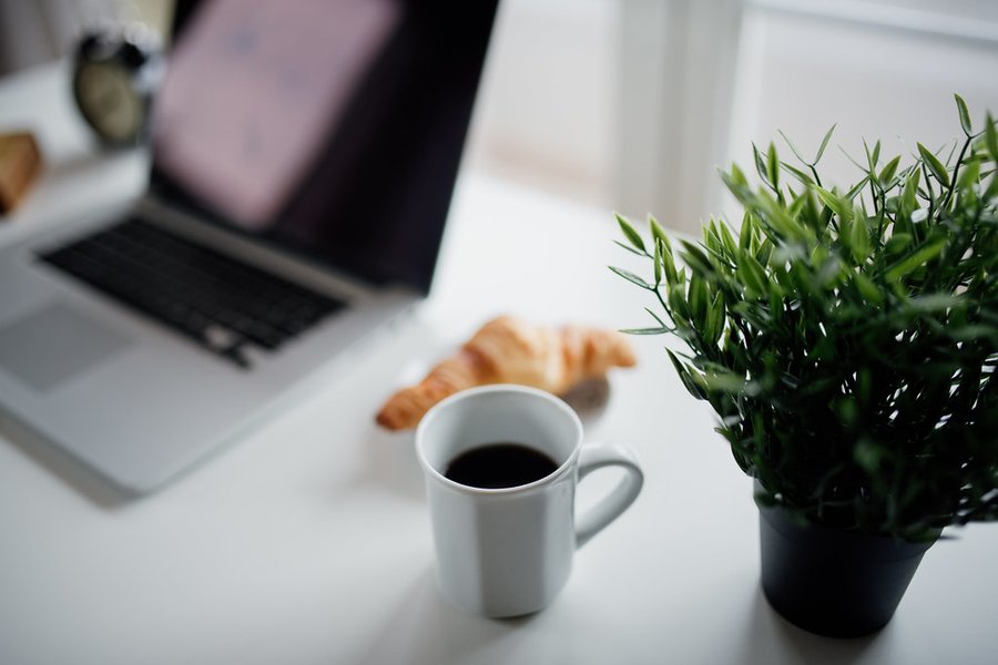 Lyst skrivebord med bærbar PC, kaffekopp, croissant og grønn plante